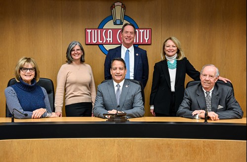 Photo of three women and three men. A woman is on the left sitting, next to two men sitting. Behind them from left to right is a woman, man, and a woman. The Tulsa County logo is behind the group on a wood panelled wall.